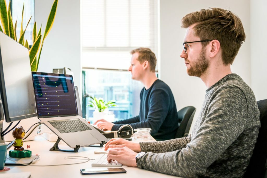 a person sitting at a desk with a laptop and another man sitting at him