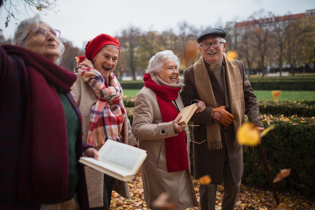 Group of happy senior friends with books on walk outdoors in park in autumn, talking and laughing
