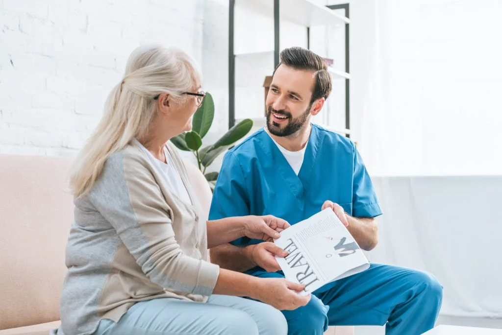 smiling male nurse looking at senior woman in eyeglasses holding travel newspaper