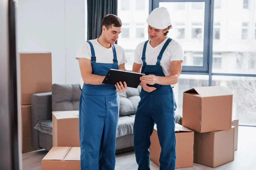 Holds notepad with document. Two young movers in blue uniform working indoors in the room