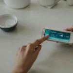 a person placing a small, portable blood testing device on a kitchen counter next to a smartphone app displaying health statistics.